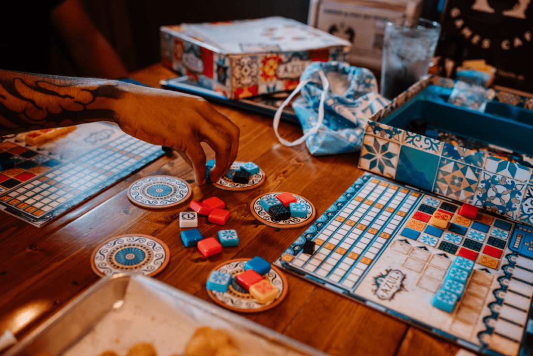 Photo zoomed in of someone's hand with a tattoo on their arm. The person is about to pick up red and blue board game pieces from a table indoors at Unplugged Games Cafe. On the table are board games that are blue, white, orange and red. 