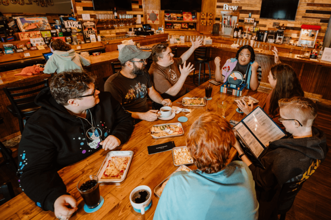 Photo taken from indoors at Unplugged Games Cafe. Seven people sit around a table with food and drinks, all while enjoying a board game. One person is looking at a menu. Theres a wooden bar in the background with one patron sitting at it.