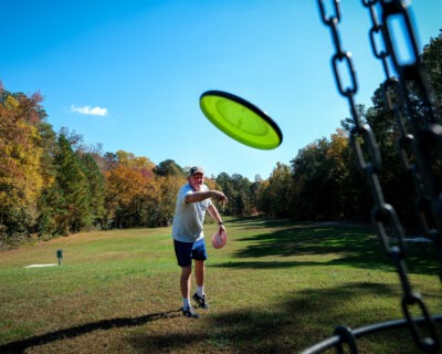 man throwing green disc towards the disc hole. He is standing on green grass with green trees in the background.
