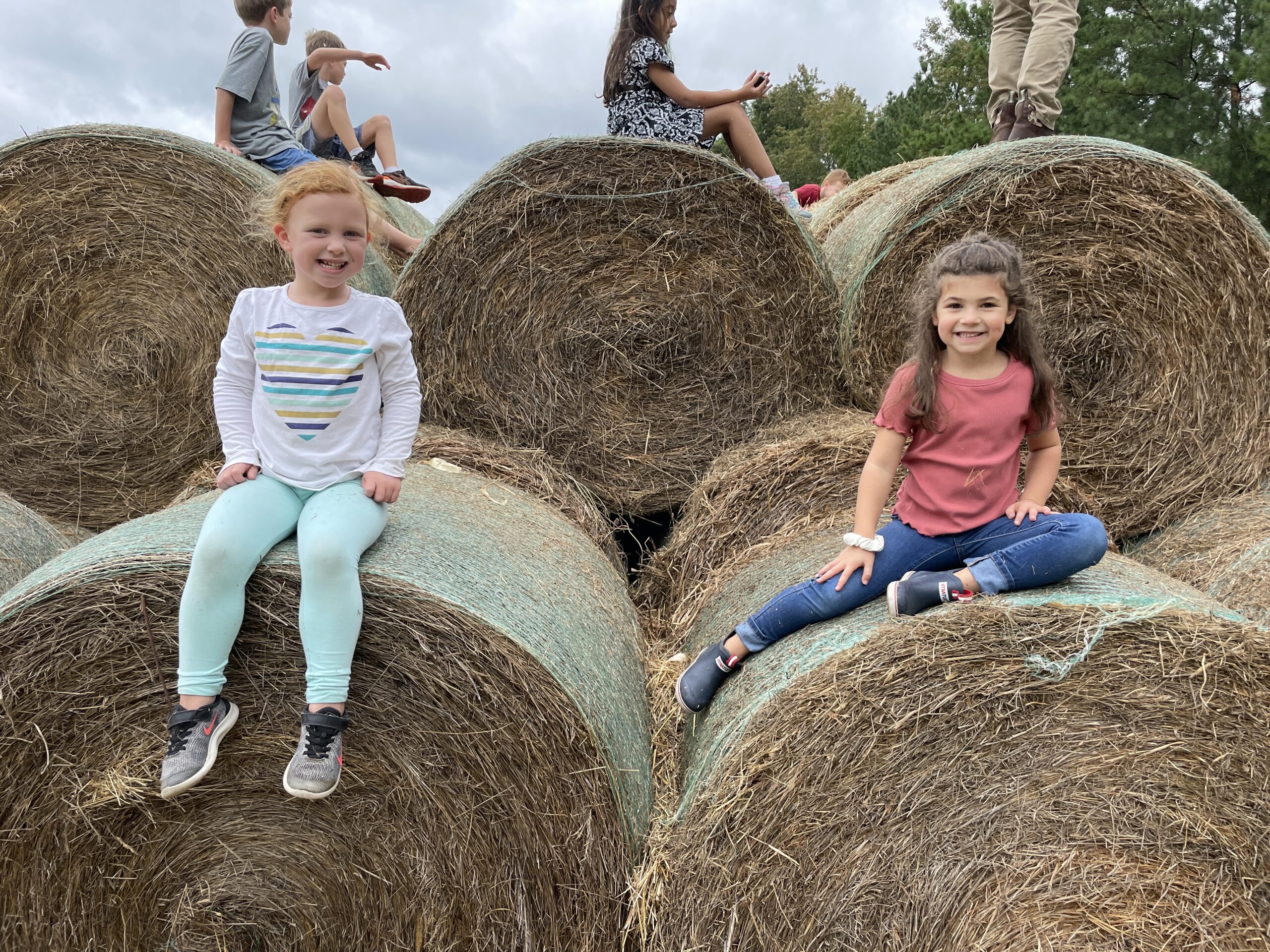 Two little girls sit upon hay bales at Skinquarter Farm. One is wearing blue tights with a white shirt and red hair. The other is wearing jeans, boots, a burnt red shirt and brown hair. They are both smiling.