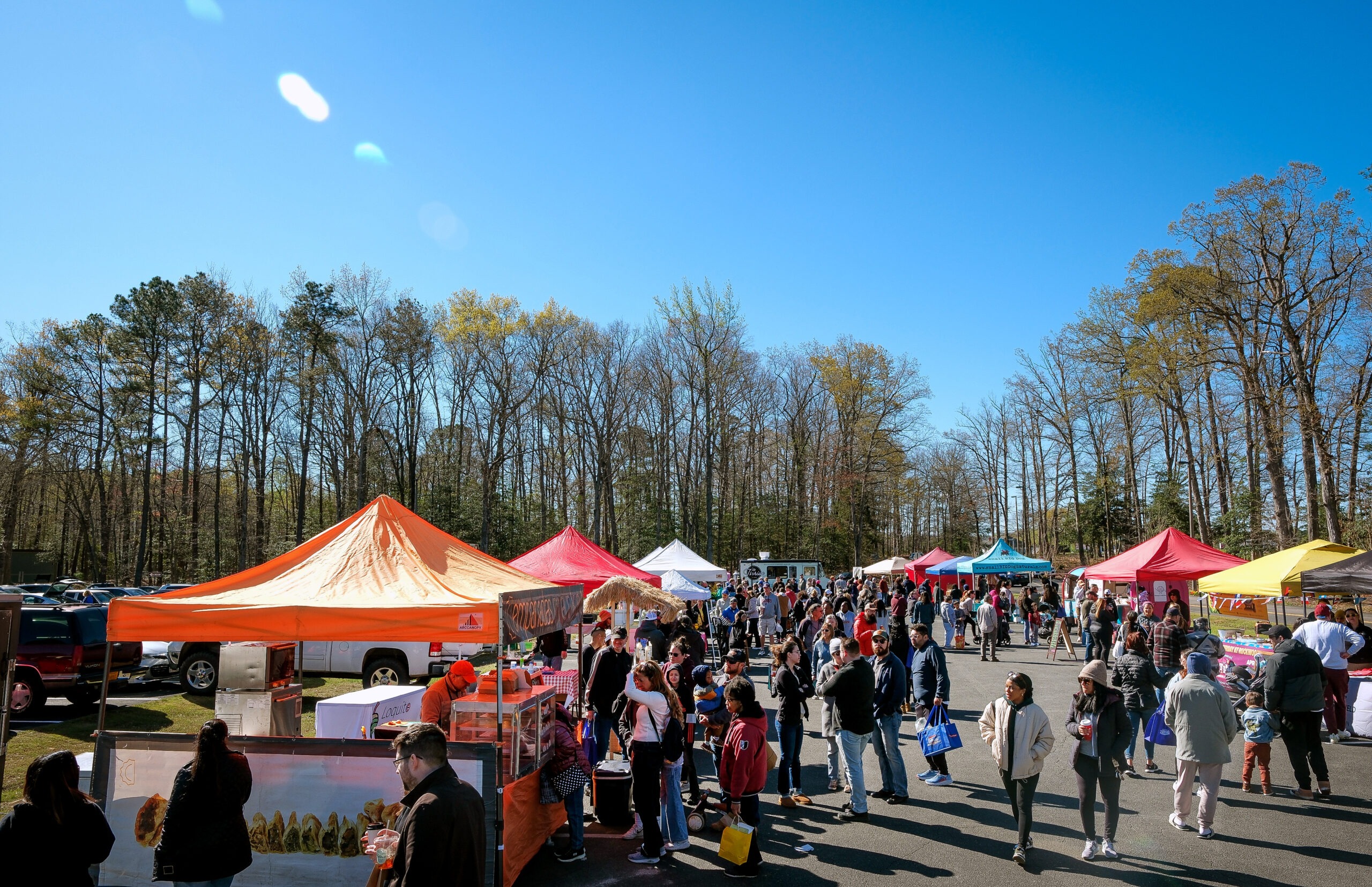 colorful tents set up on left and right side of photo with hundreds of people in the middle, shopping from the vendors under the tents. Tents are colorful: red, orange, white, blue and yellow. Most people are wearing sweatshirts, jackets, sunglasses and boots on this chilly day. Trees in background are losing their leaves. 